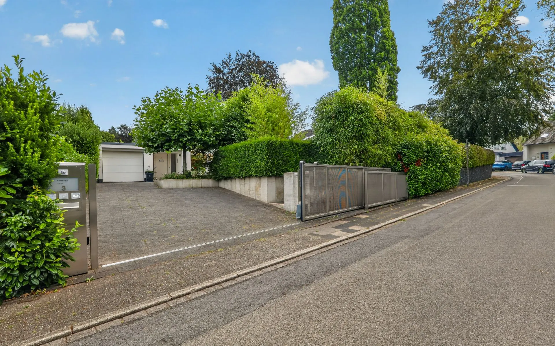 Einfahrt zu einem Wohnhaus mit Metalltor, Auffahrt und viel grüner Bepflanzung.