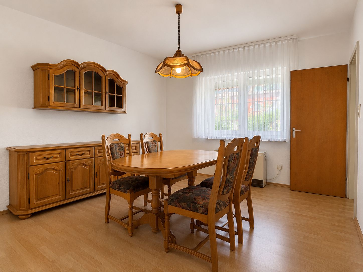 Esszimmer mit Holztisch, vier Stühlen, Vitrine, Kommode und Fenster mit Gardinen.