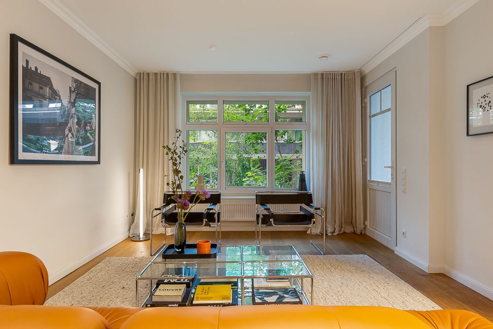 Helles Wohnzimmer mit gelbem Sofa, Glastisch, zwei Stühlen und großem Fenster mit Vorhängen.