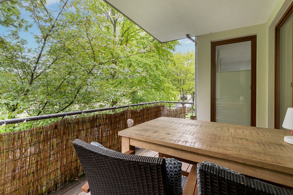 Überdachter Balkon mit Holztisch, zwei Stühlen und Ausblick auf grüne Bäume.