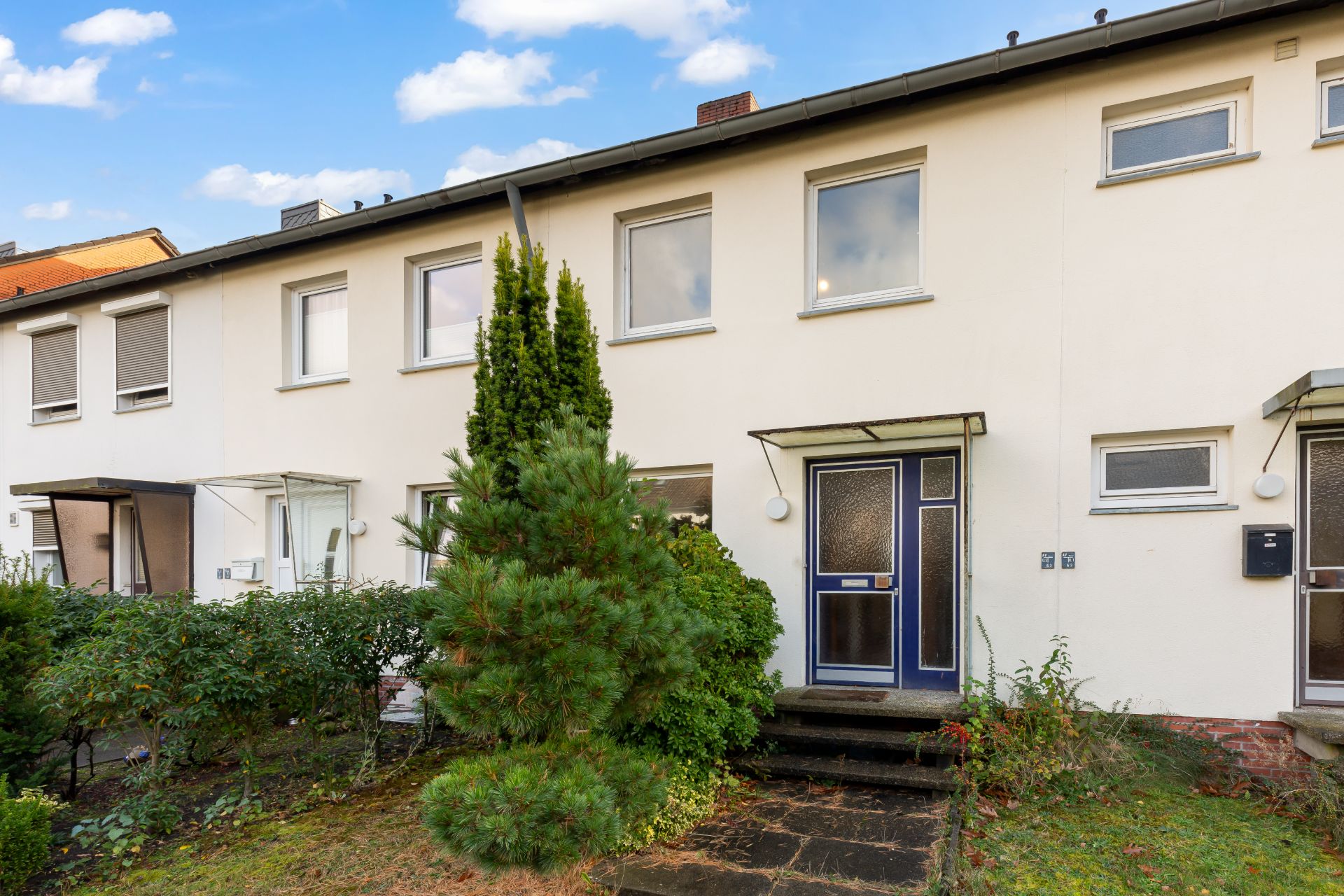 Reihenhaus mit weißer Fassade, großem Fenster und kleinem Garten mit Sträuchern und Baum vorne.