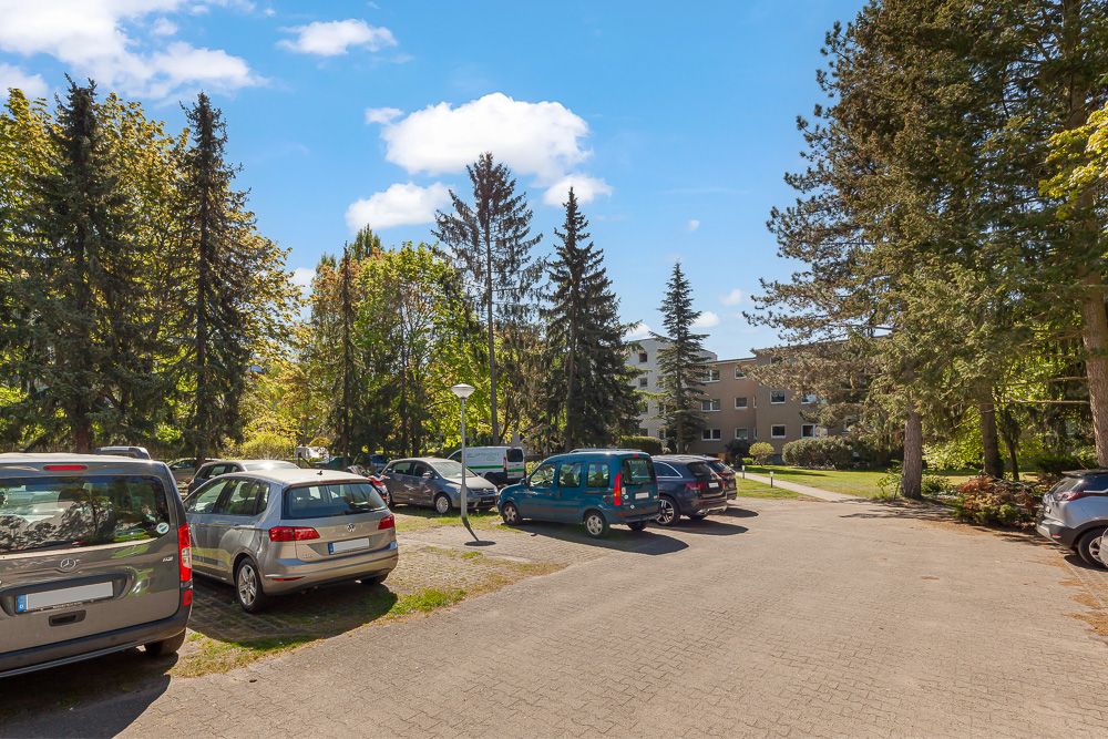 Parkplatz mit mehreren Autos, umgeben von Bäumen, Wohngebäude im Hintergrund bei sonnigem Wetter.