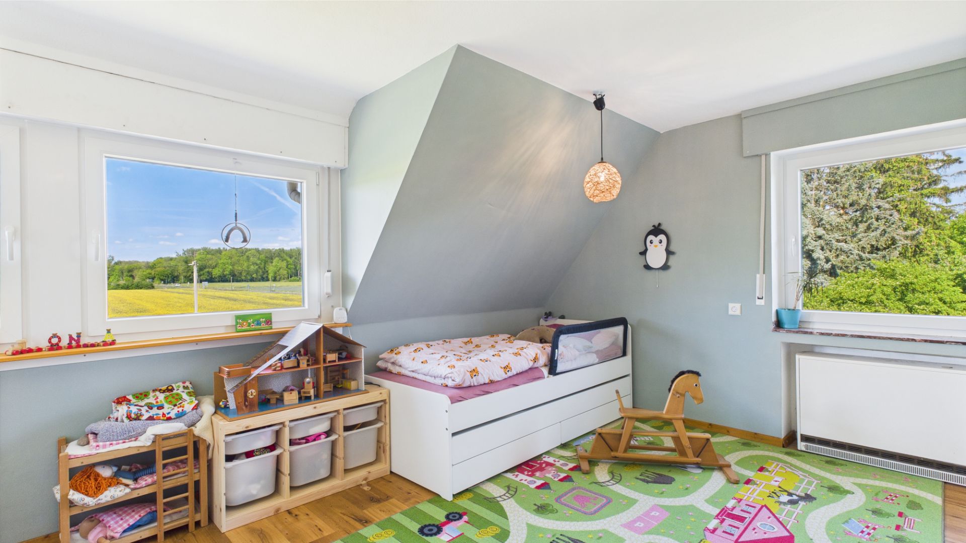 Helles Kinderzimmer mit Bett, Spielzeugregal, Schaukelpferd und buntem Teppich, zwei große Fenster.