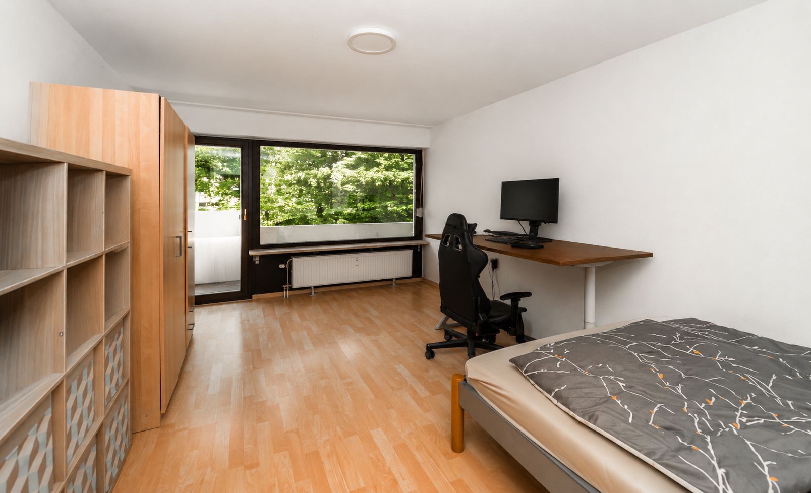 Helles Zimmer mit Bett, Schreibtisch, Stuhl, Regal, Kleiderschrank und großem Fenster.