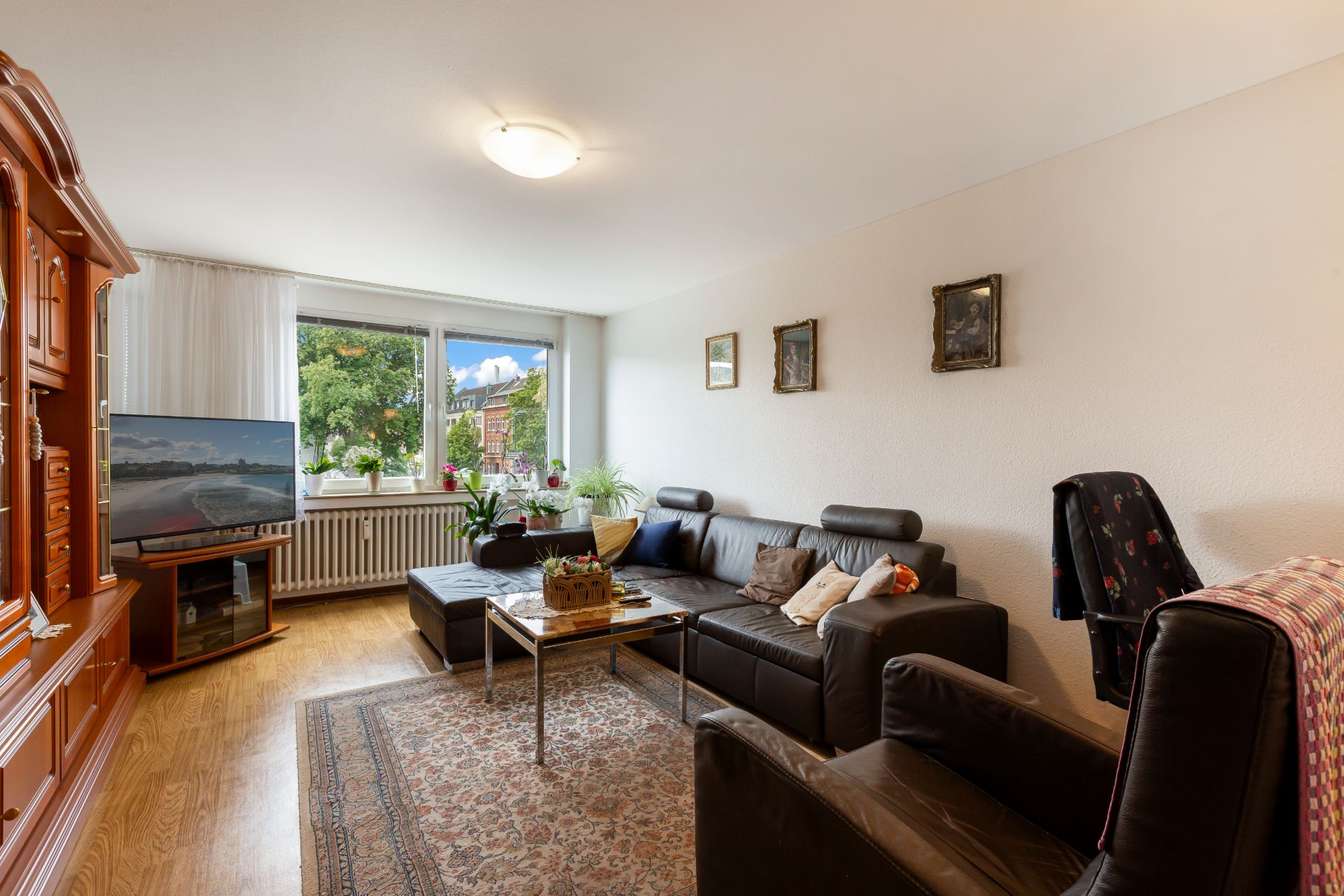 Helles Wohnzimmer mit brauner Ledercouch, Teppich, Holzmöbeln und großem Fenster mit Blick ins Grüne.