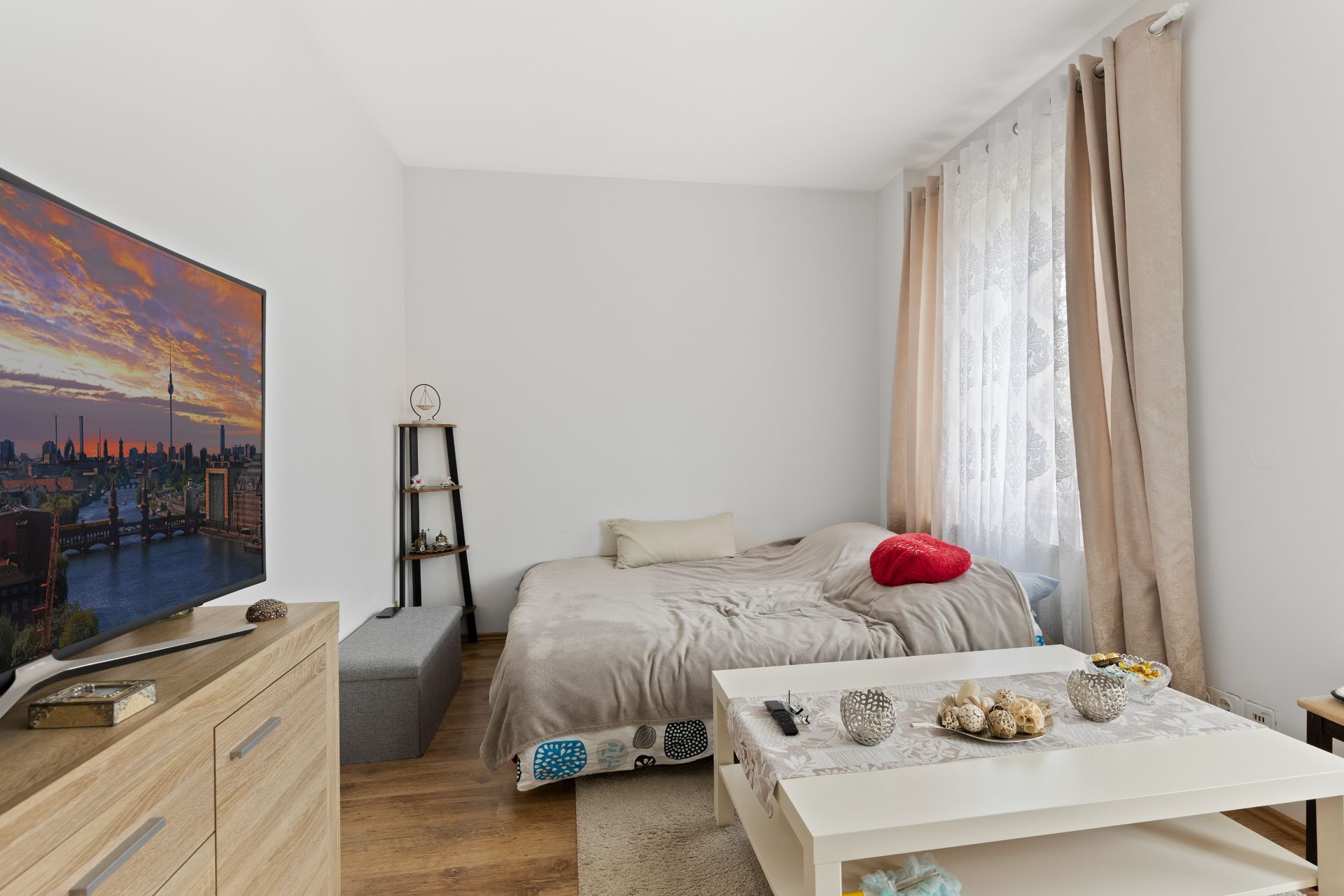 Helles Schlafzimmer mit Bett, Couchtisch, Fernseher und großem Fenster mit Vorhängen.