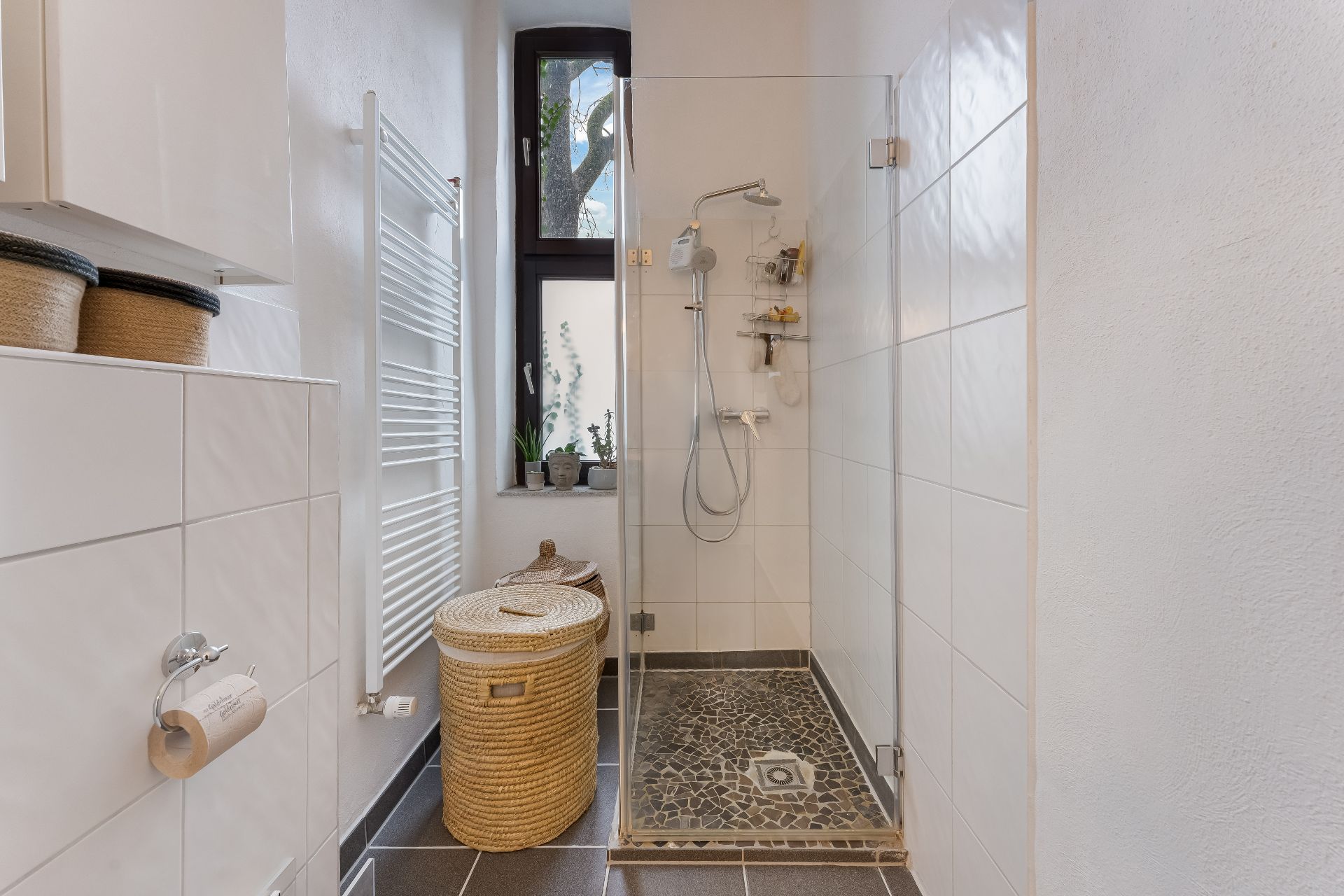 Helles Badezimmer mit bodengleicher Dusche, Fenster, Heizkörper und Wäschekorb.