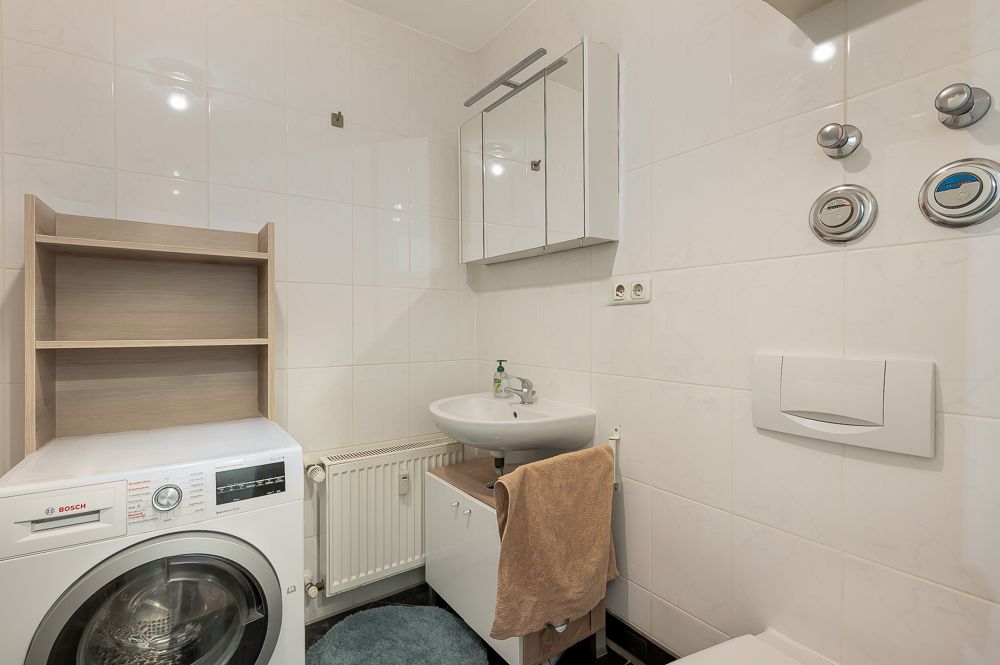Kleines Bad mit Waschbecken, Waschmaschine, Regal, Spiegel und Wandtoilette.