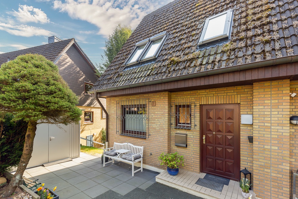 Eingang eines Einfamilienhauses mit brauner Tür, Bank und Baum im Vorgarten.