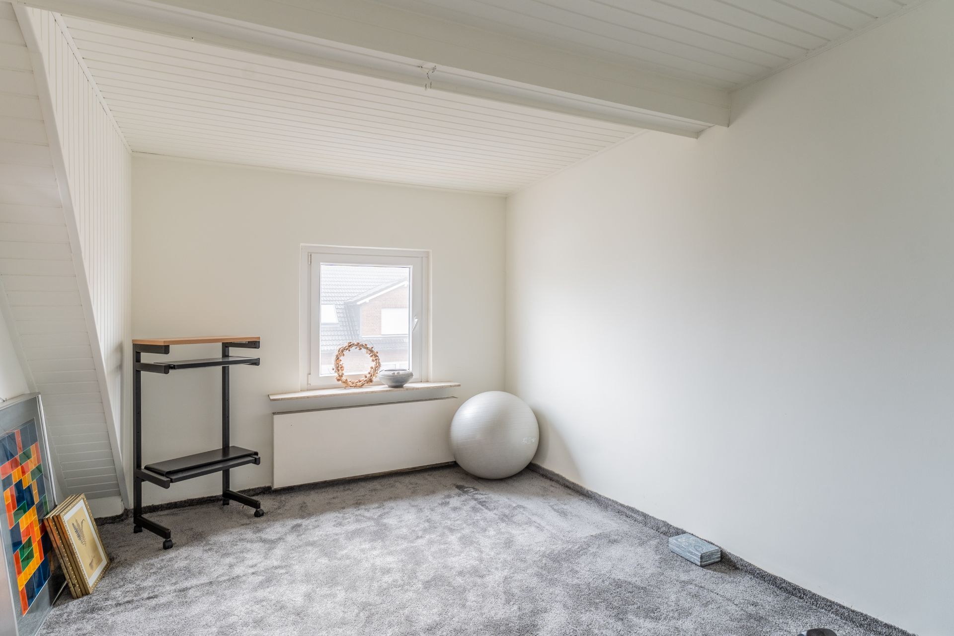 Heller Raum mit grauem Teppich, Fenster, Gymnastikball und kleinem Regal.