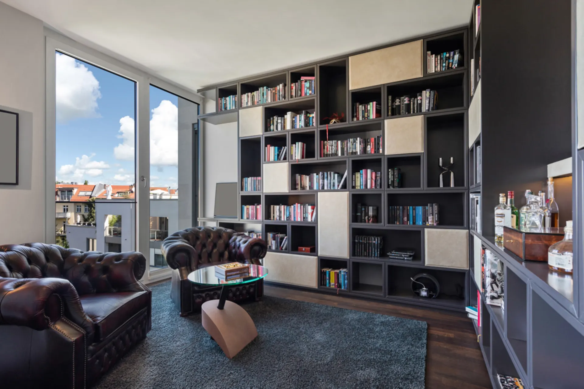 Wohnzimmer mit großen Bücherregalen, zwei Ledersesseln, Glastisch und Balkonblick.