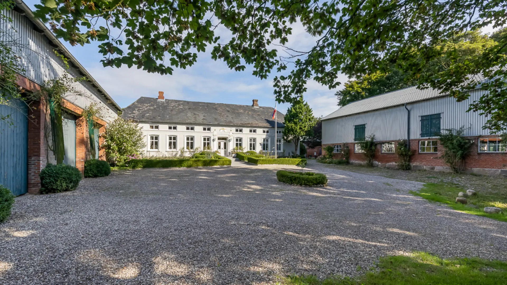 Hof mit Kiesfläche, zentralem weißen Wohnhaus und zwei Nebengebäuden, umgeben von Bäumen.
