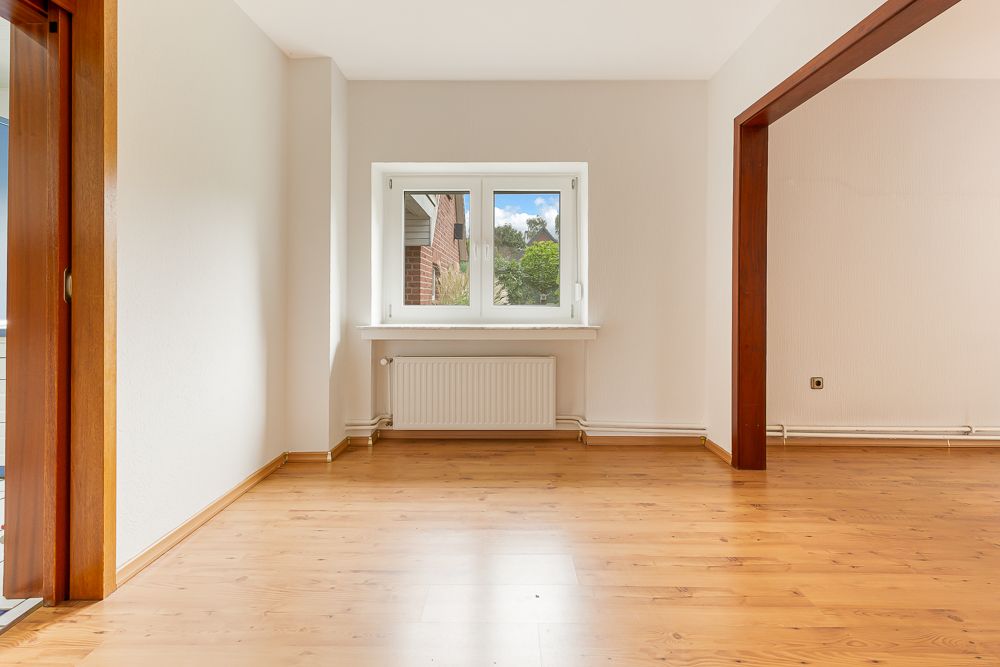 Leerer Raum mit Holzfußboden, weißer Wand und Fenster mit Blick ins Grüne.