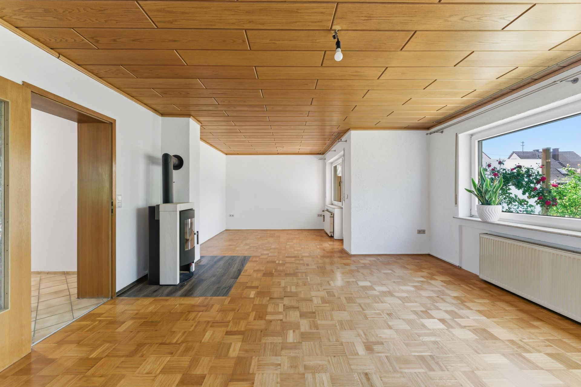Helles Wohnzimmer mit Parkettboden, Holzdecke, Kaminofen und großem Fenster mit Pflanzen.
