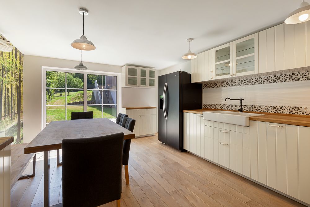 Helle Küche mit Holzboden, Esstisch, schwarzen Stühlen und großem Fenster mit Blick ins Grüne.