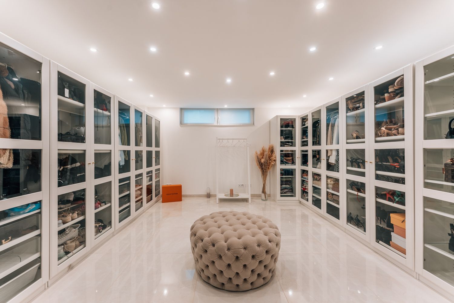 Großer, moderner Ankleideraum mit weißen Schränken und rundem, gepolstertem Sitz in der Mitte.