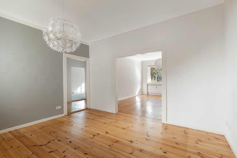 Helles Zimmer mit Holzboden, grauer und weißer Wand, großer Lampe und Durchgang zu weiterem Raum.