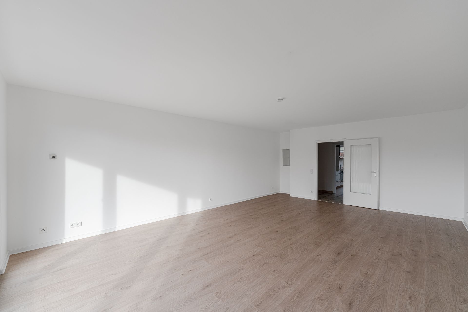 Leerer, heller Raum mit Holzfußboden und weißen Wänden, Tür zum nächsten Zimmer sichtbar.