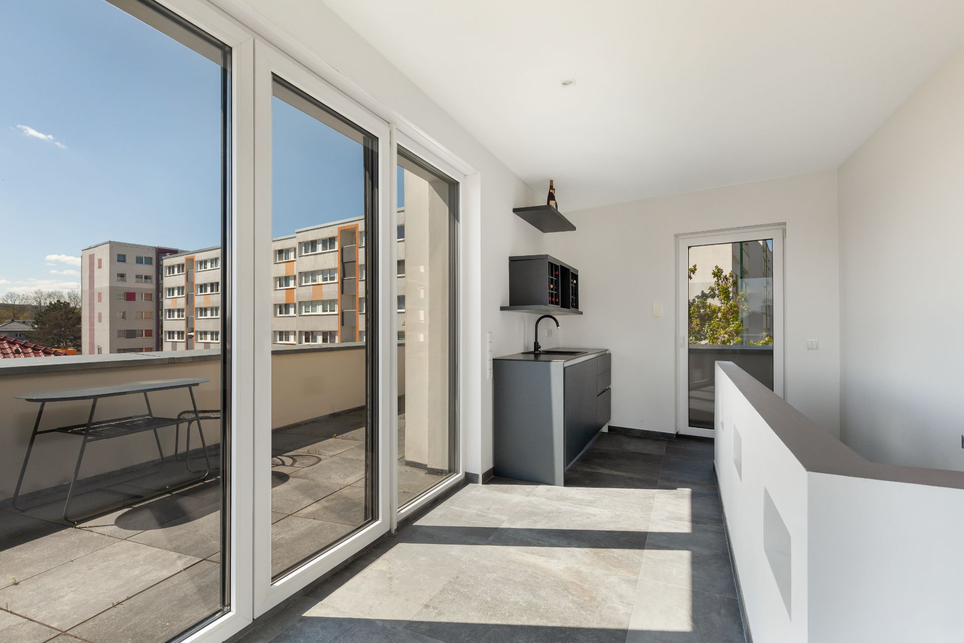 Moderner Balkon mit Glas-Schiebetüren, kleiner Küchenzeile und Blick auf benachbarte Wohngebäude.