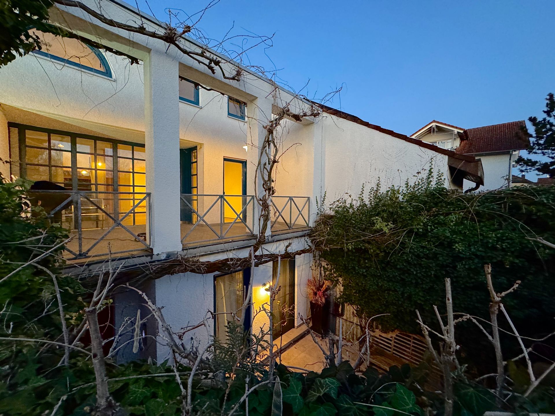 Zweistöckiges Wohnhaus mit Balkon, großer Fensterfront und Garten im Vordergrund bei Abenddämmerung.