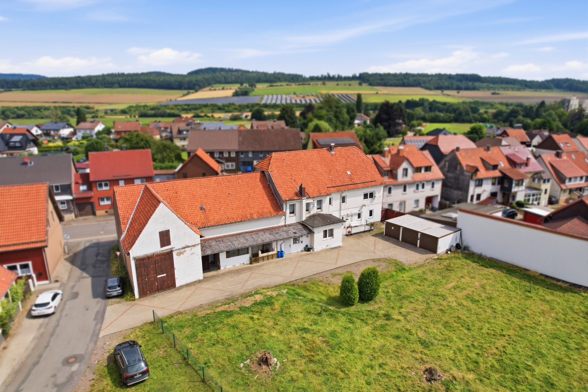 Luftaufnahme eines Wohnhauses mit rotem Dach, Hof und angrenzender Wiese in einem Dorf.