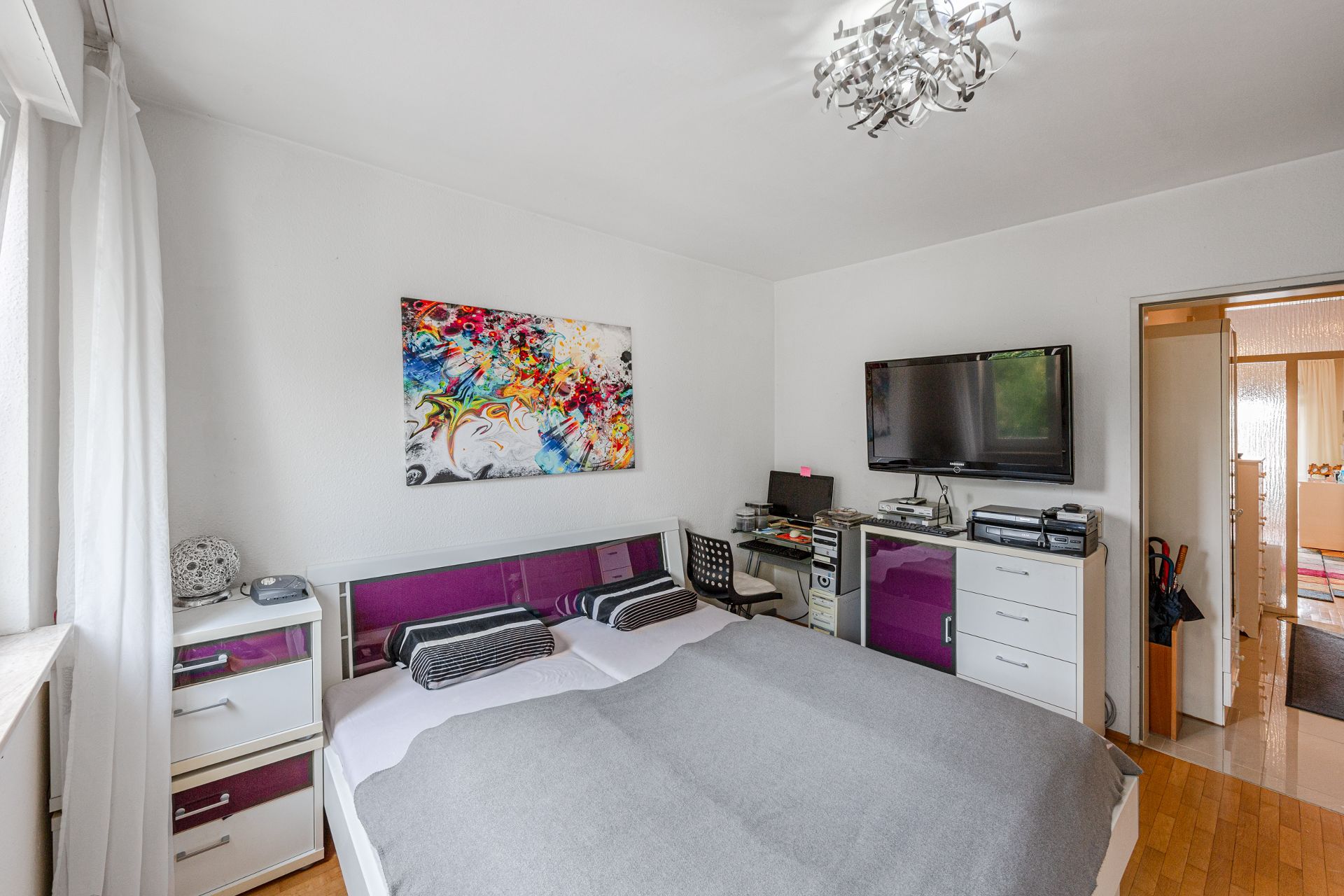 Schlafzimmer mit Doppelbett, bunter Wandkunst, Schreibtisch, Fernseher und Kommode.