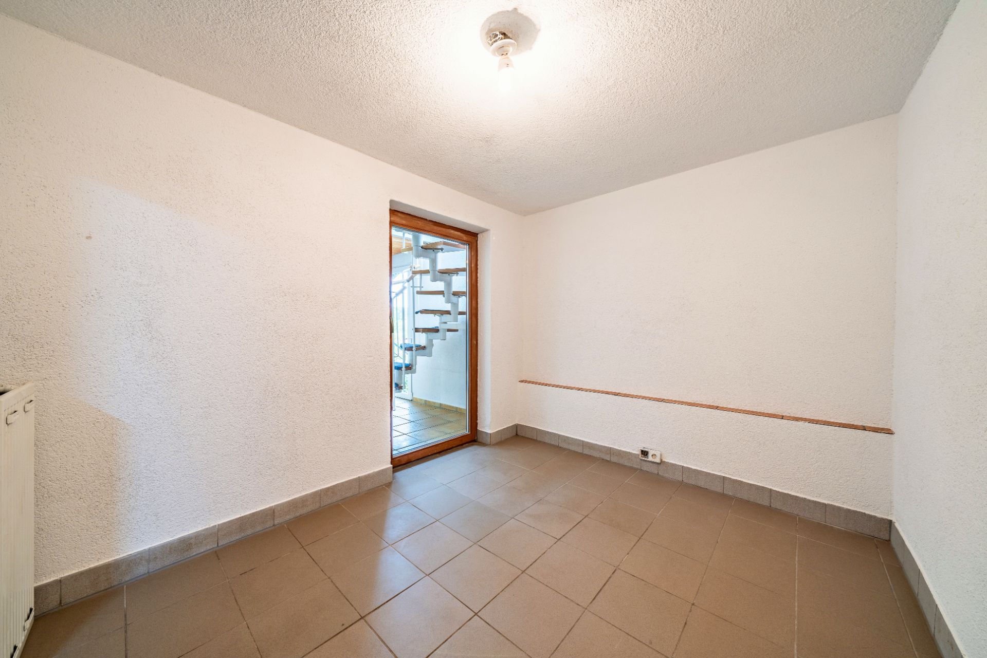 Leerer Raum mit braunen Fliesen, weißen Wänden und Tür zu einer Treppe.