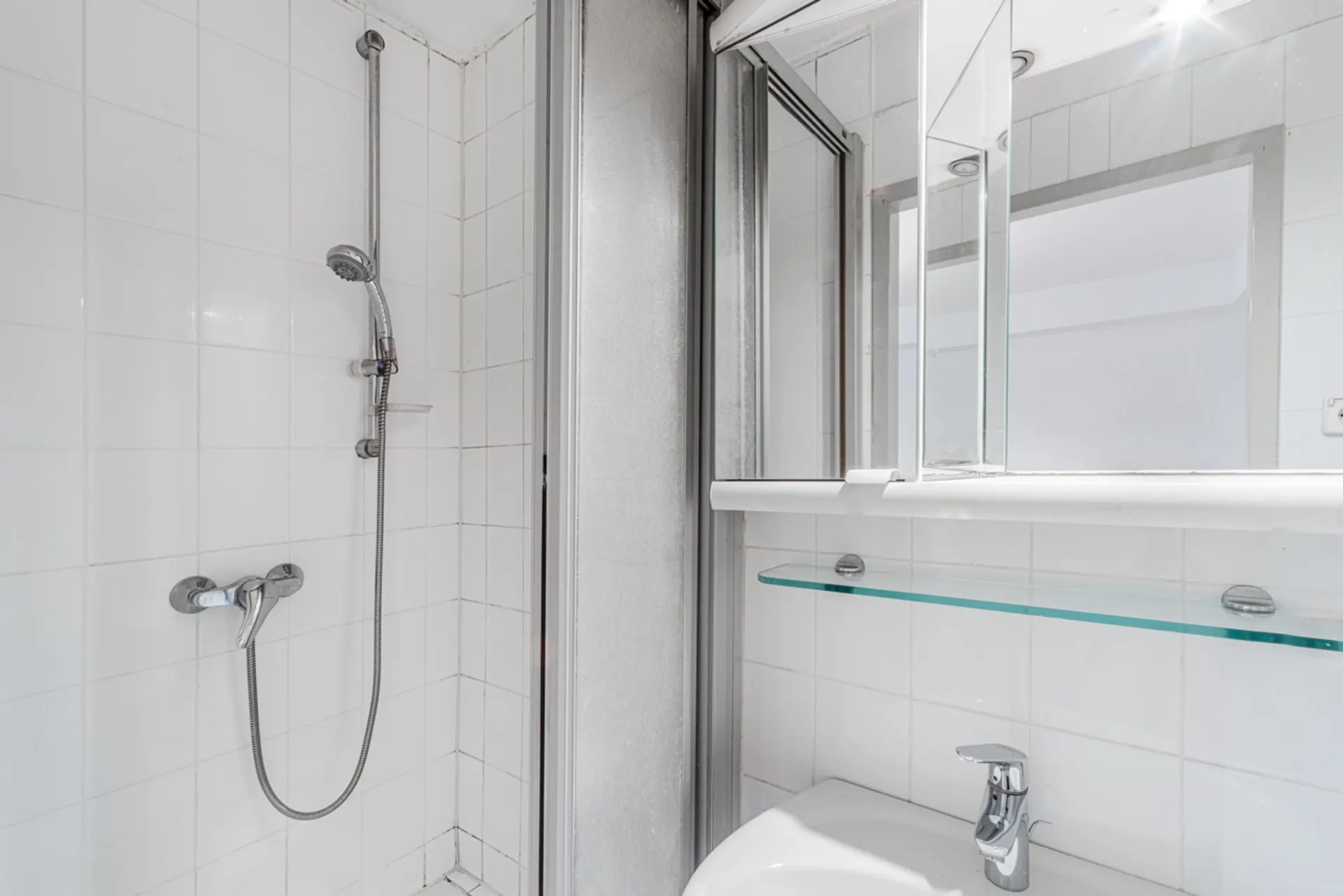 Dusche mit Glaswand, Waschbecken, Glasablage und Spiegel in einem weißen Badezimmer.