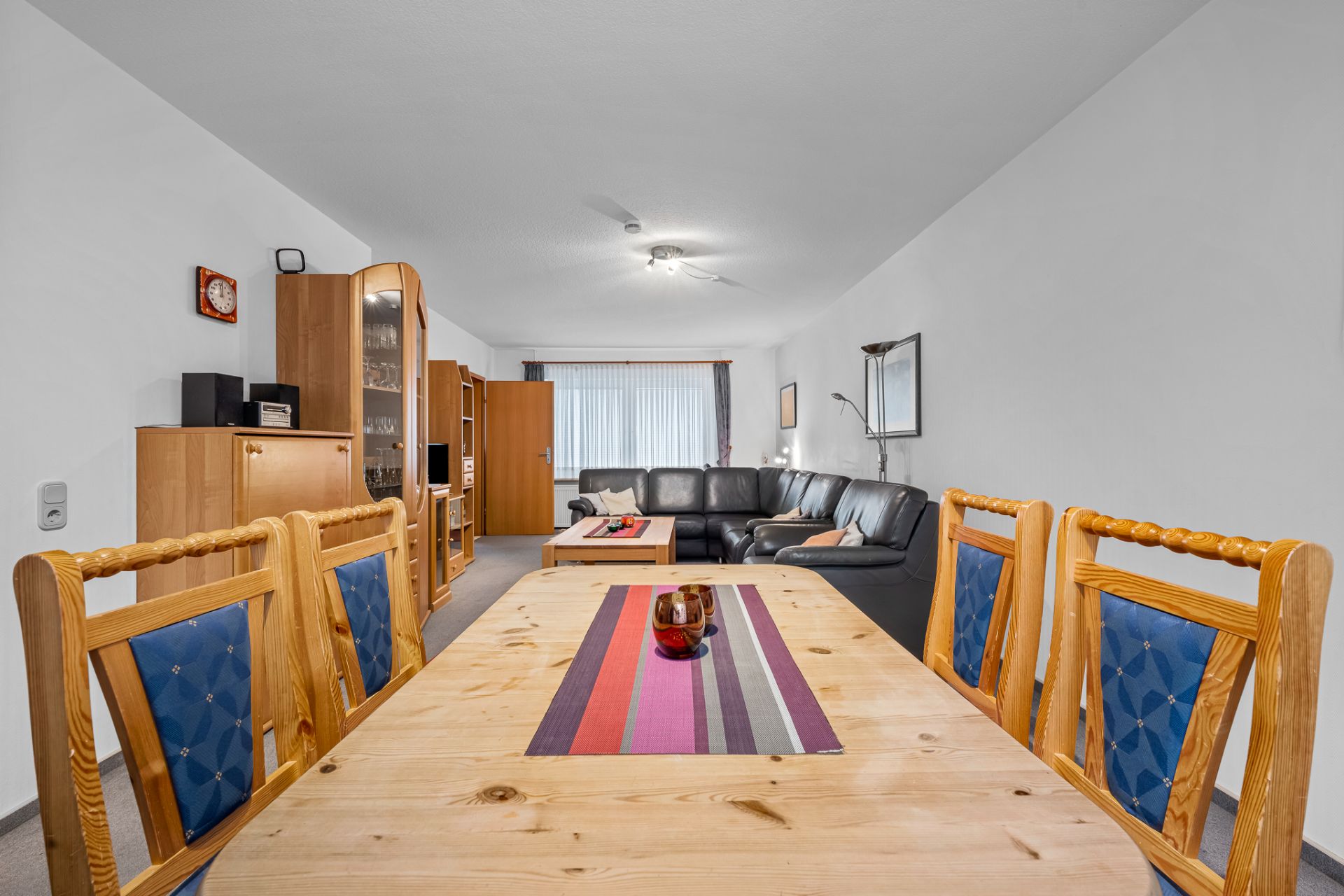 Wohnzimmer mit Holz-Esstisch, Stühlen, schwarzem Sofa und Schrankwand.