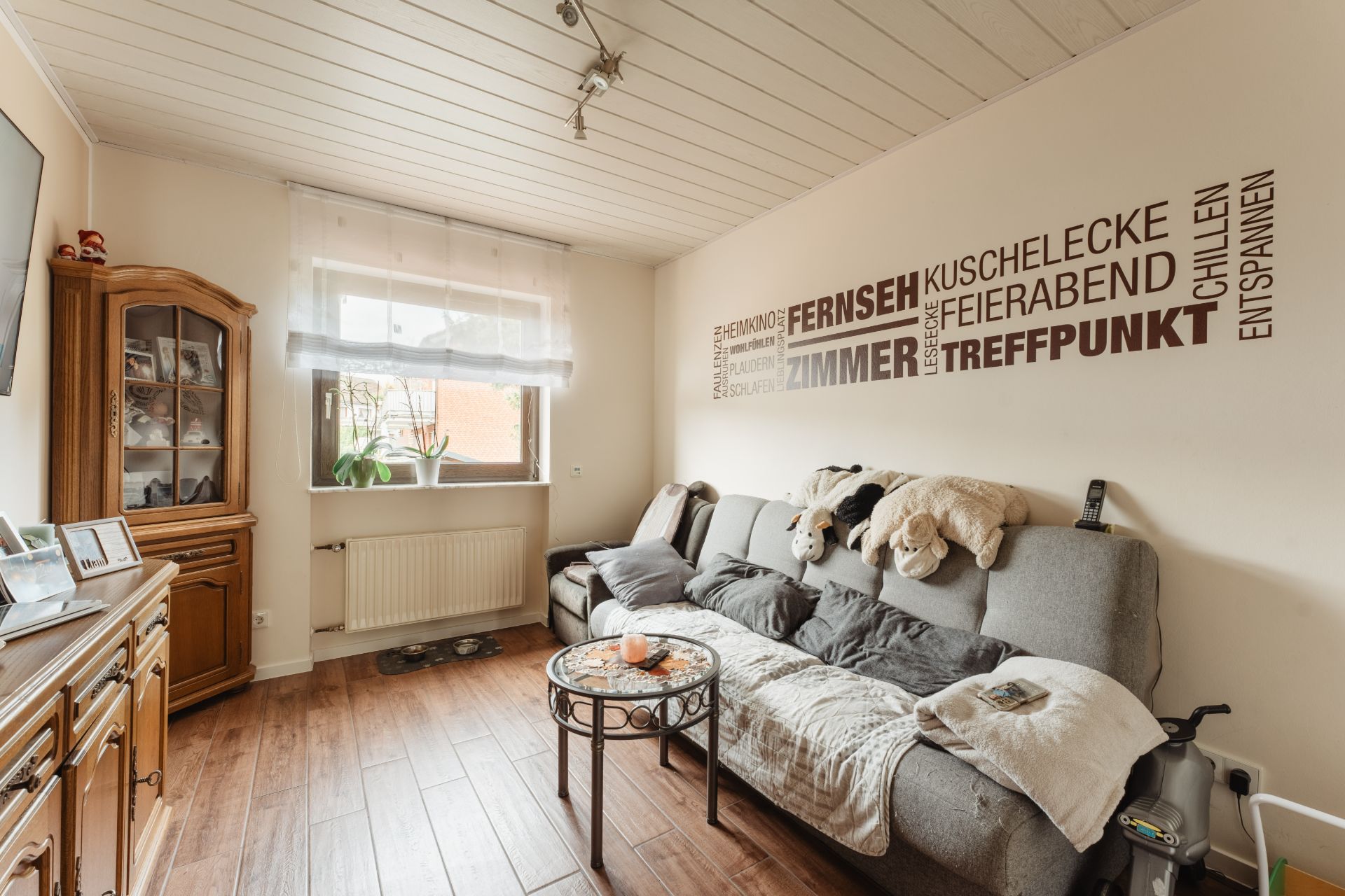 Gemütliches Wohnzimmer mit grauem Sofa, Holzmöbeln und Wanddeko mit Schriftzügen.