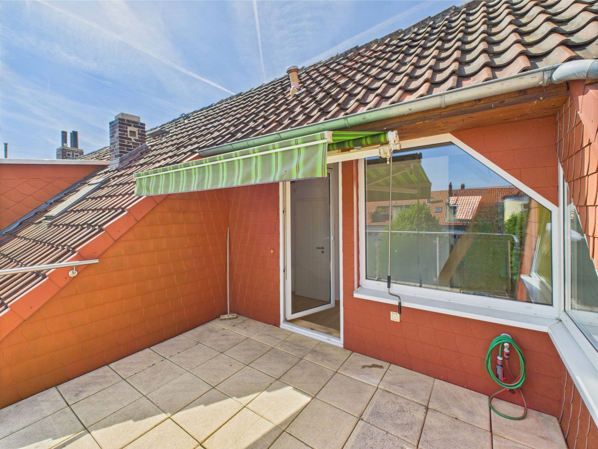 Dachterrasse mit Fliesenboden, Markise und Zugang durch eine Tür, Fenster und Gartenschlauch an der Wand.