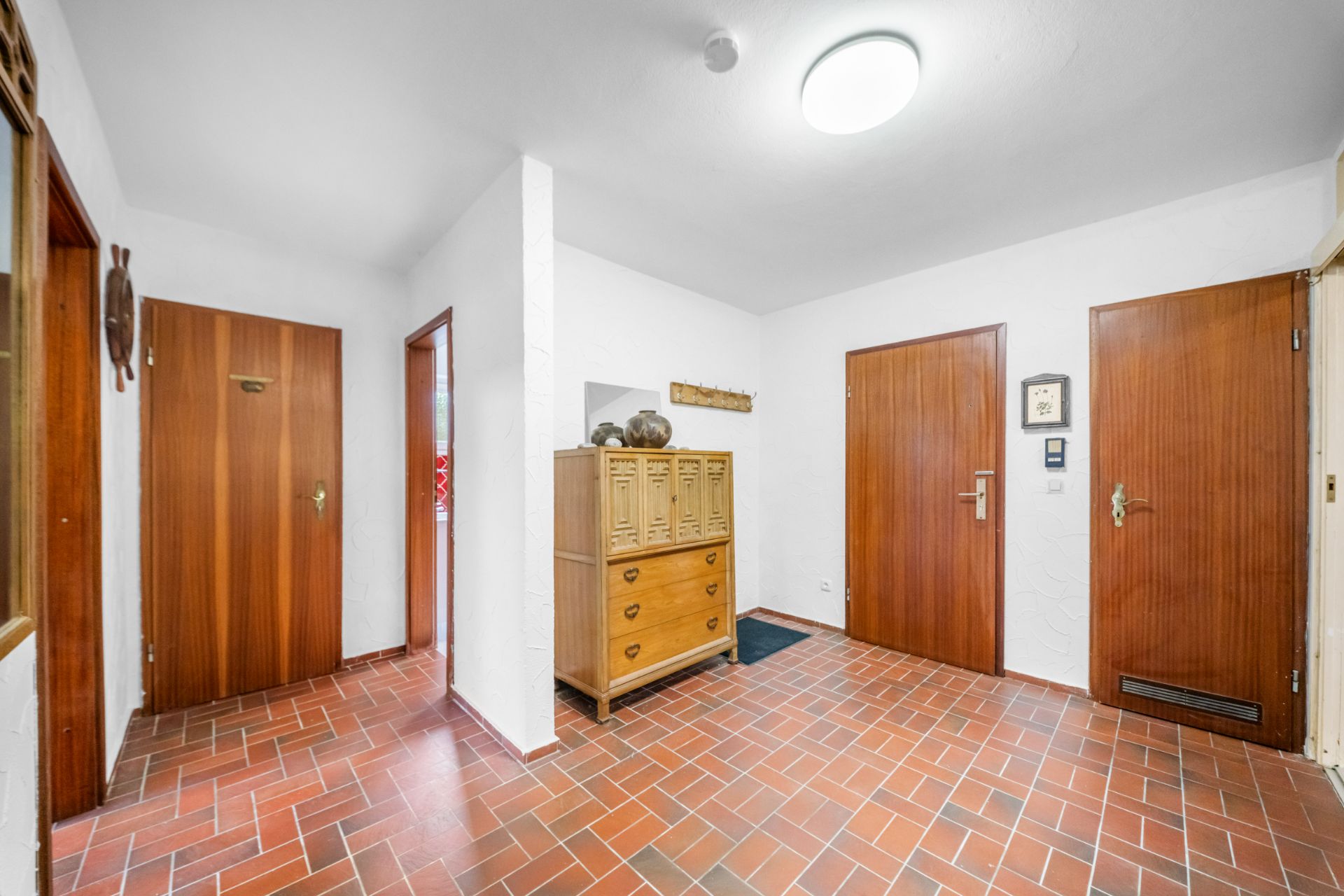 Flur mit roten Bodenfliesen, mehreren Holztüren und einer Kommode vor weißer Wand.