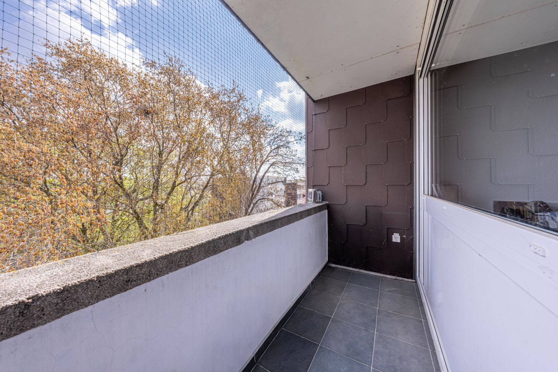 Offener Balkon mit Fliesenboden, Netzschutz und Blick auf Bäume.