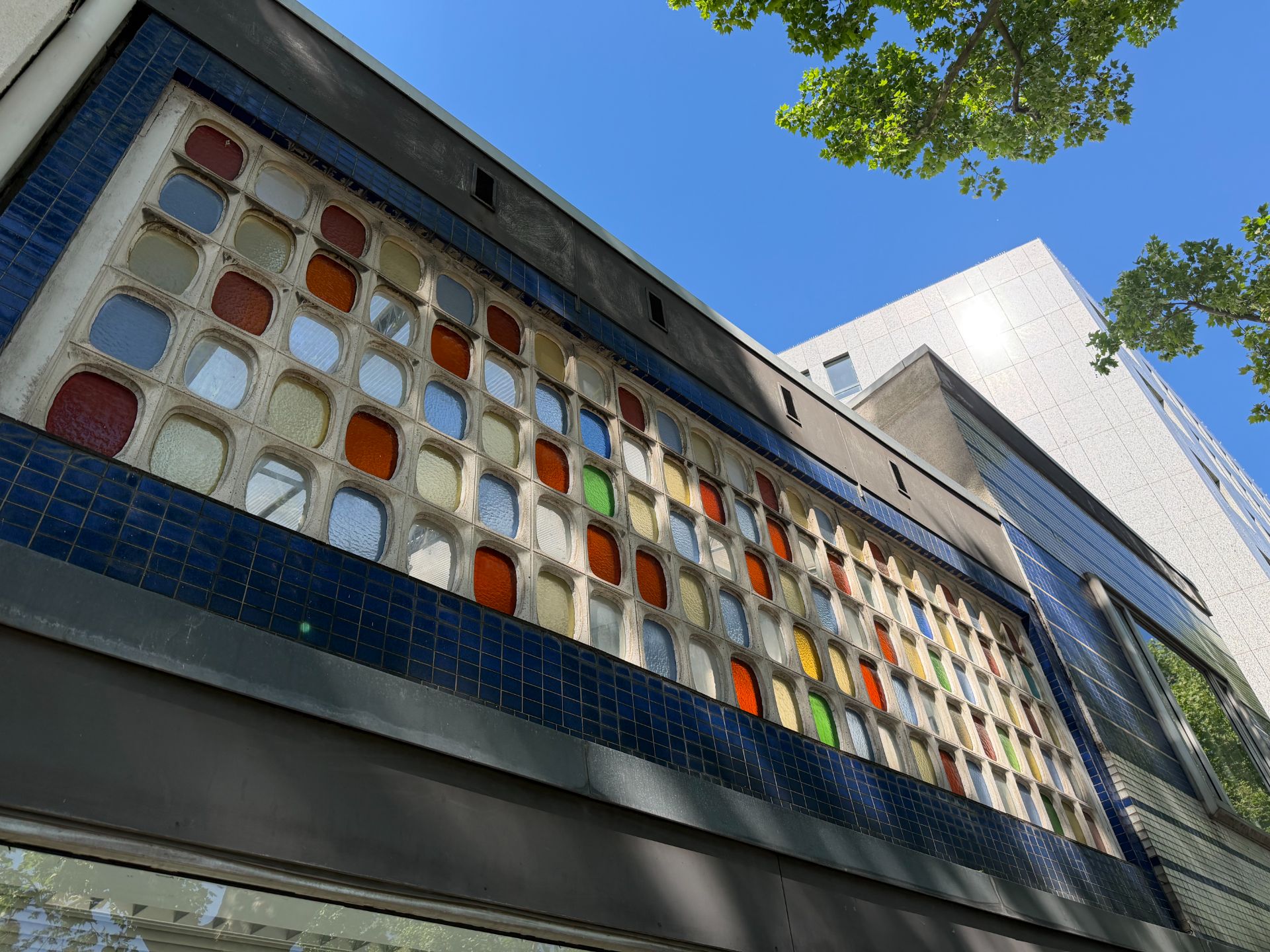 Fassade mit bunten Glasbausteinen und blauen Fliesen unter blauem Himmel und Bäumen.