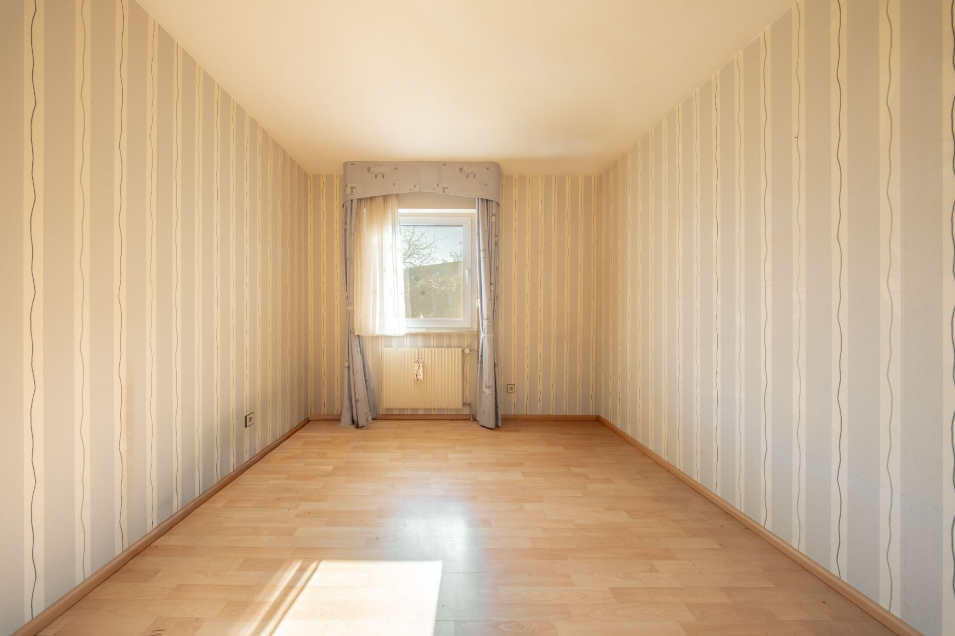 Leerer Raum mit hellem Laminatboden, gestreifter Tapete und Fenster mit Vorhang.
