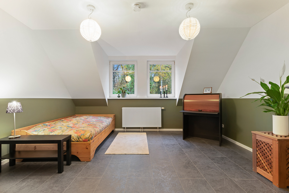 Helles Zimmer mit Einzelbett, Schreibtisch, Topfpflanze und drei Fenstern.