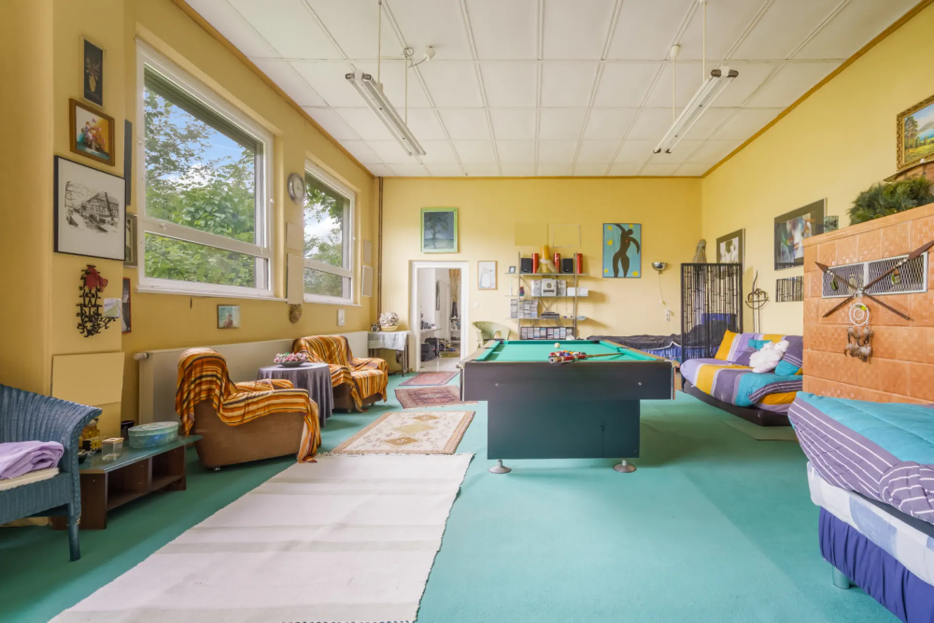 Wohnzimmer mit Billardtisch, Sofas, Sesseln und großen Fenstern, viele Wandbilder und Dekoration.