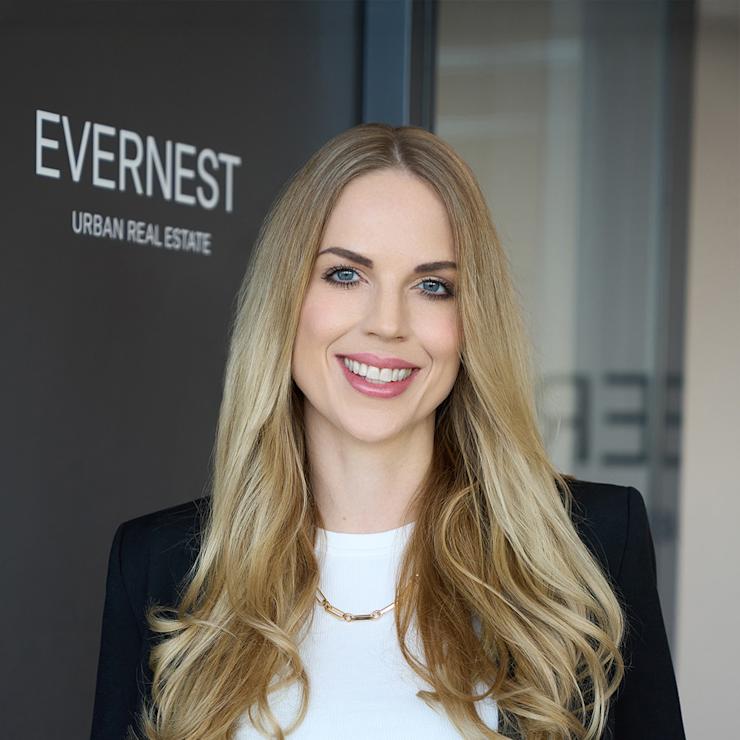Eine lächelnde Frau mit langen blonden Haaren steht vor einer Glastür mit der Aufschrift 'Evernest Urban Real Estate'.
