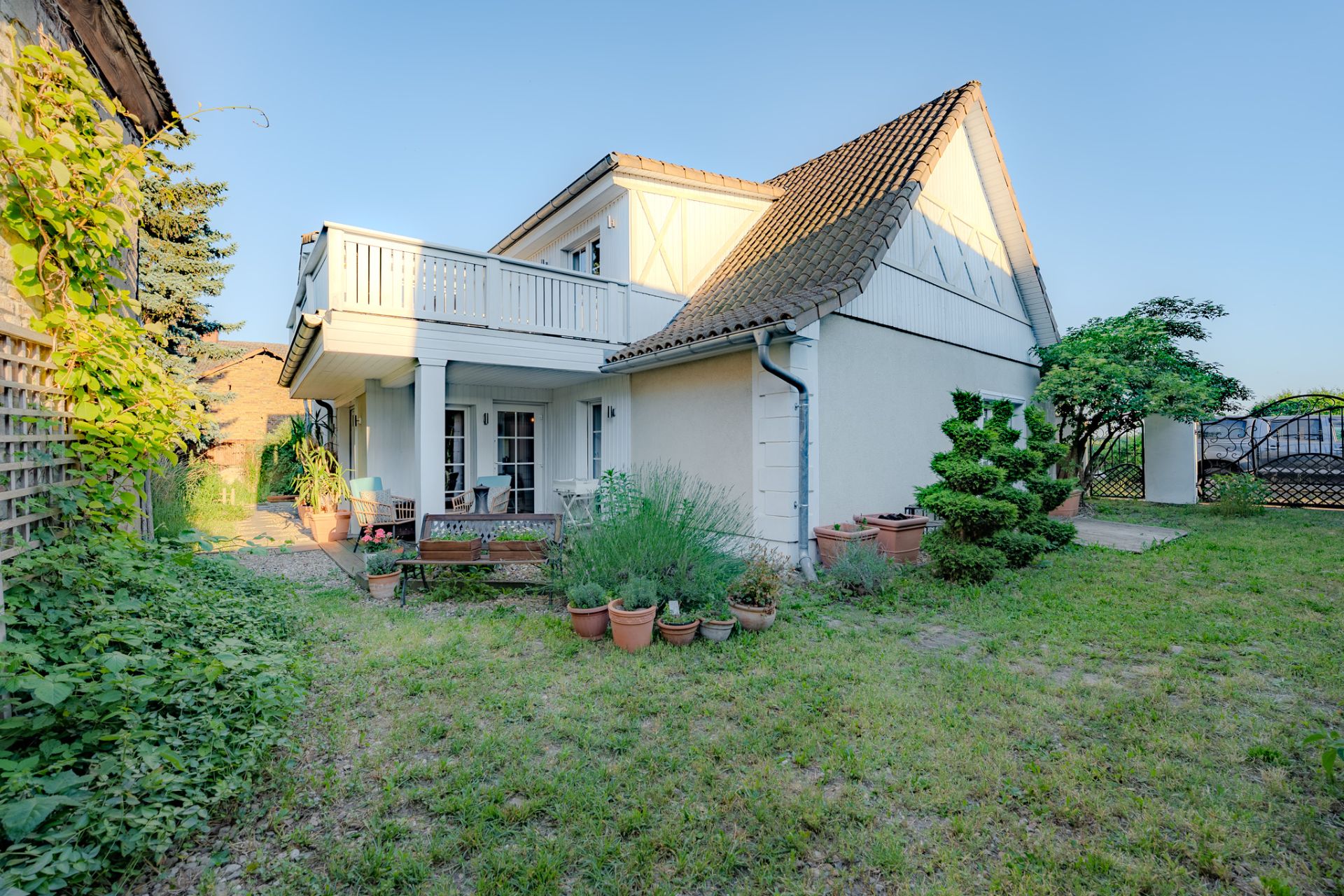 Haus mit Balkon, Terrasse, Gartenbank und vielen Blumentöpfen im Garten bei Tageslicht.