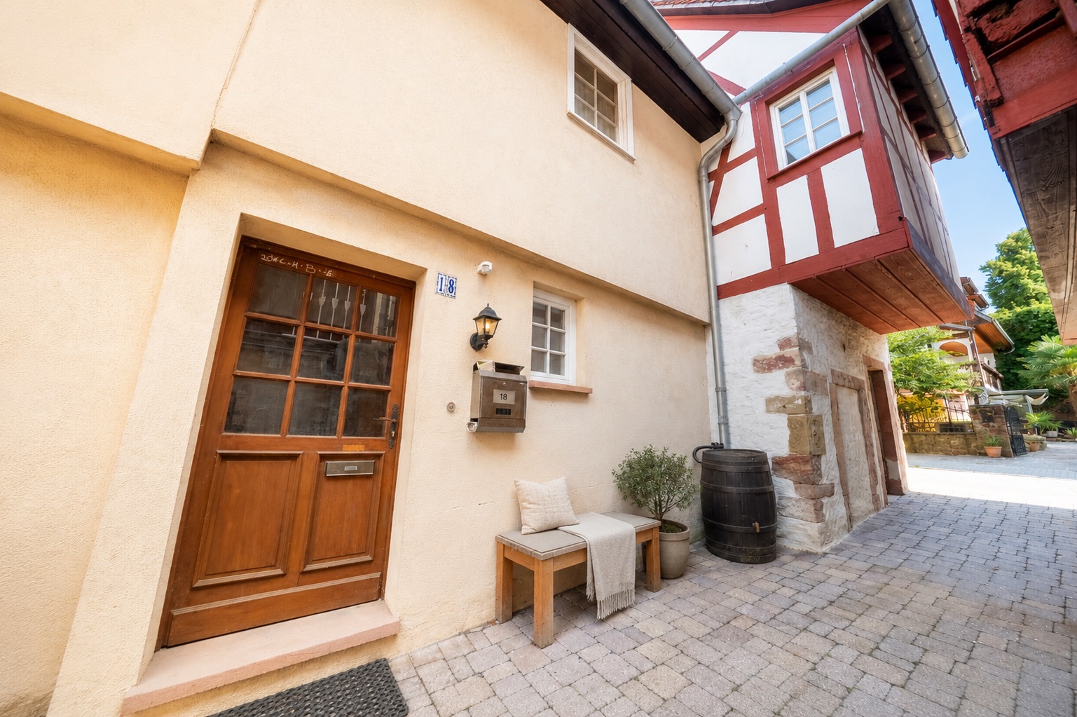 Eingangsbereich eines Hauses mit Holztür, Bank, Briefkasten und Pflanze auf Steinpflaster.