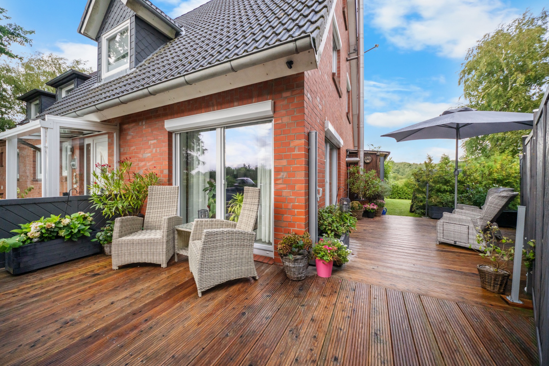 Holzterrasse an einem Wohnhaus mit Gartenmöbeln, Pflanzen und Sonnenschirm.