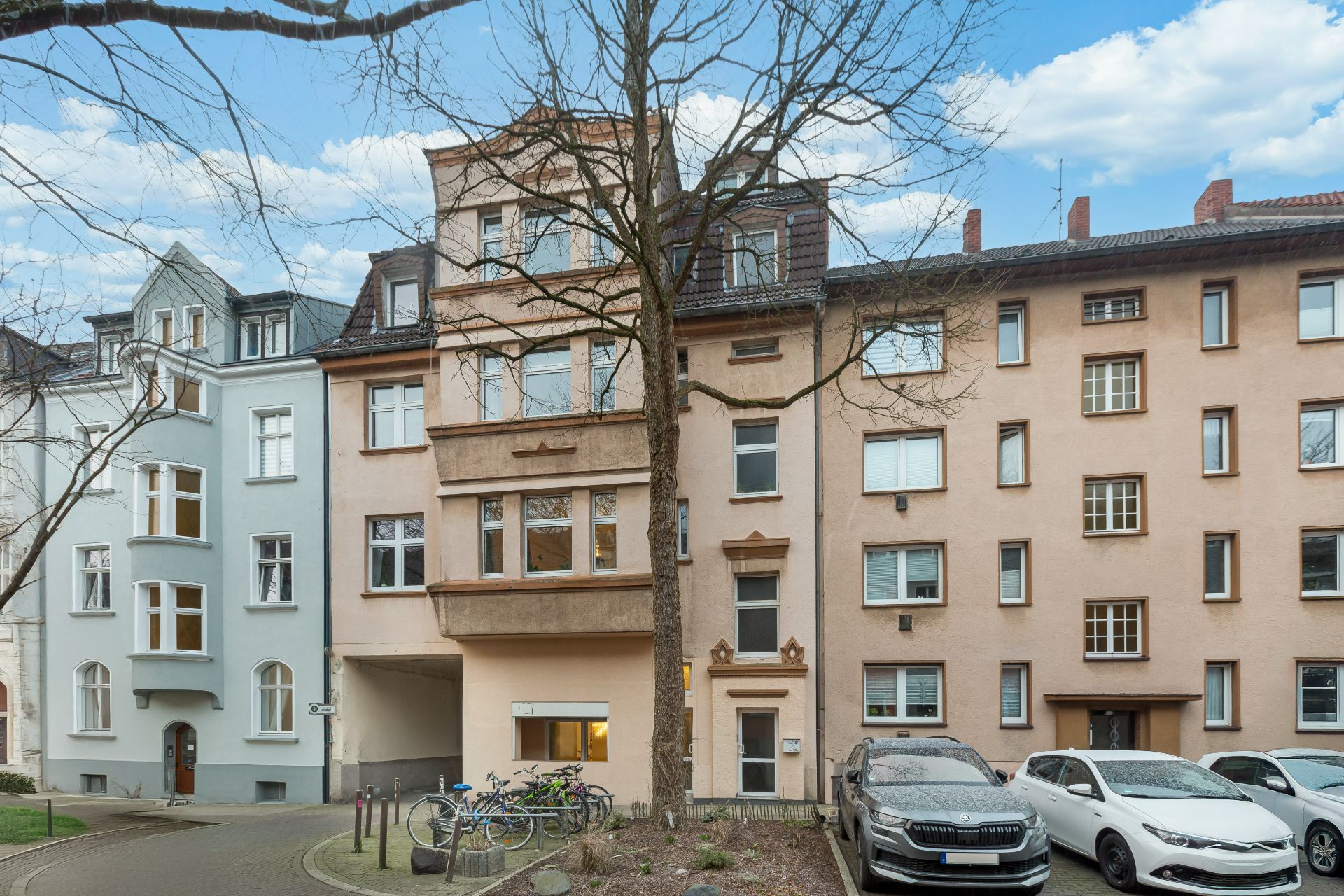Mehrstöckiges Wohnhaus mit geparkten Autos und Fahrrädern vor dem Eingang.