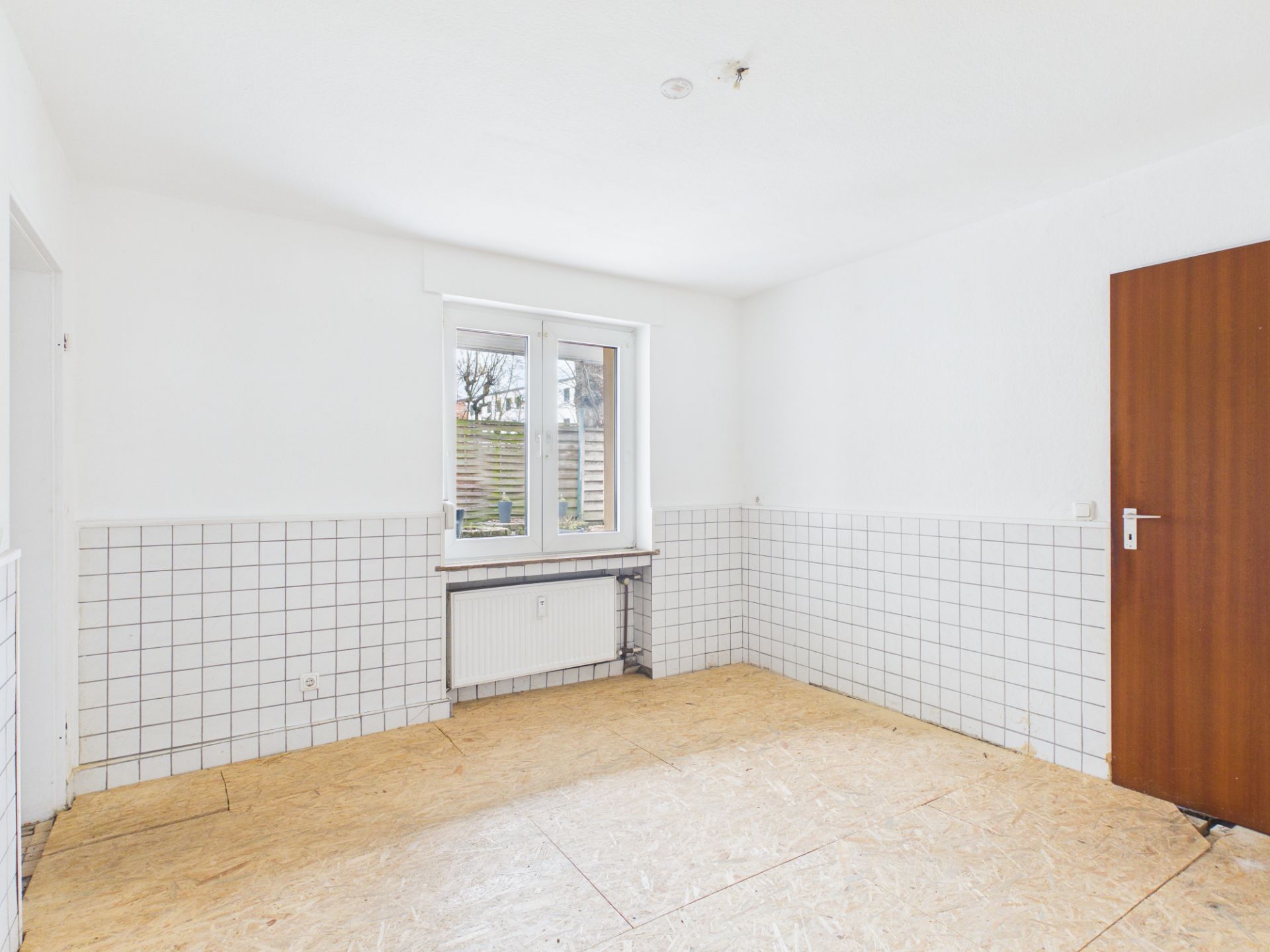 Leerer, heller Raum mit weiß gekachelten Wänden, Fenster und brauner Holztür.