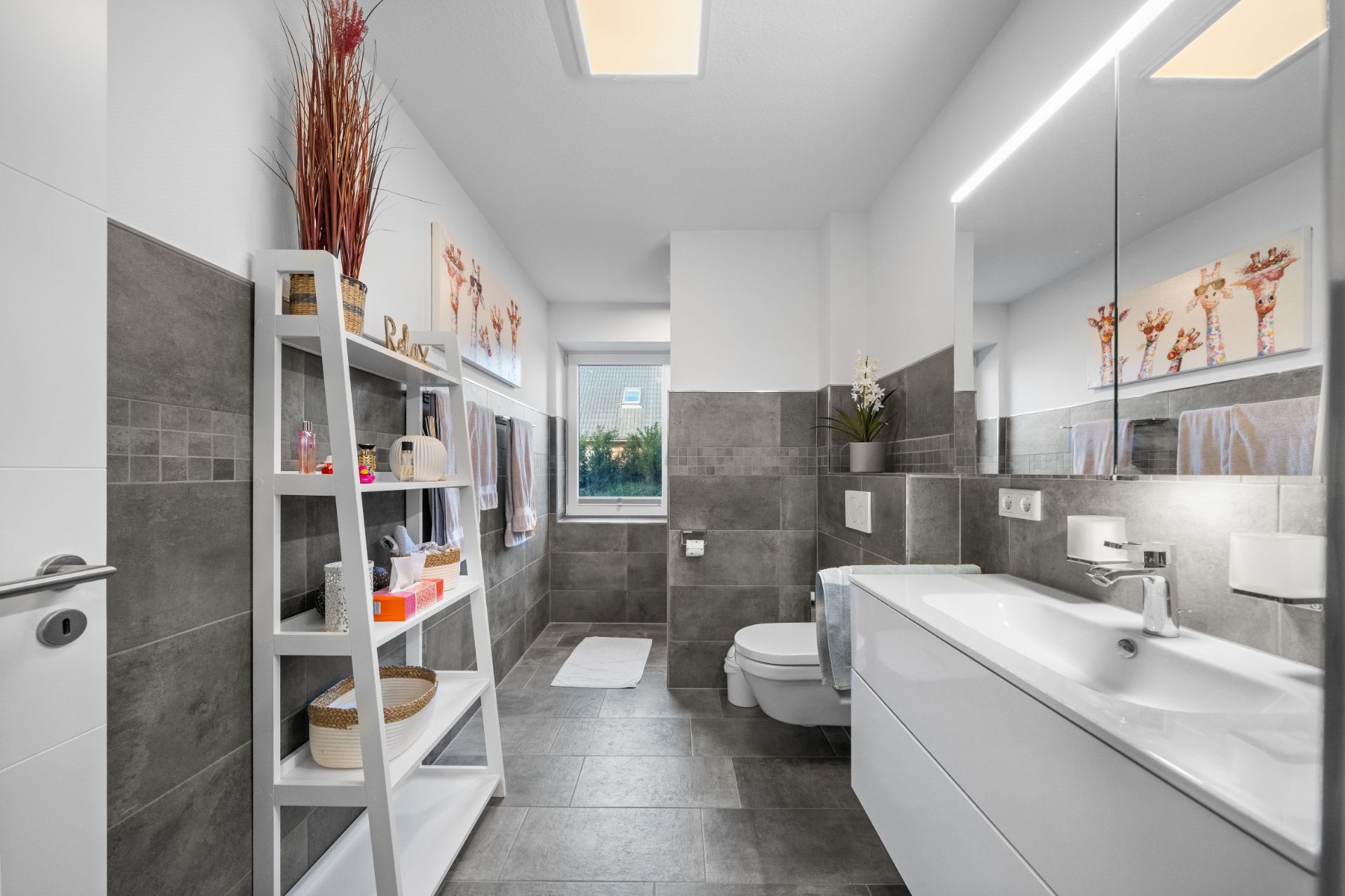 Modernes Badezimmer mit grauen Fliesen, weißer Einrichtung, Spiegel und Regal.
