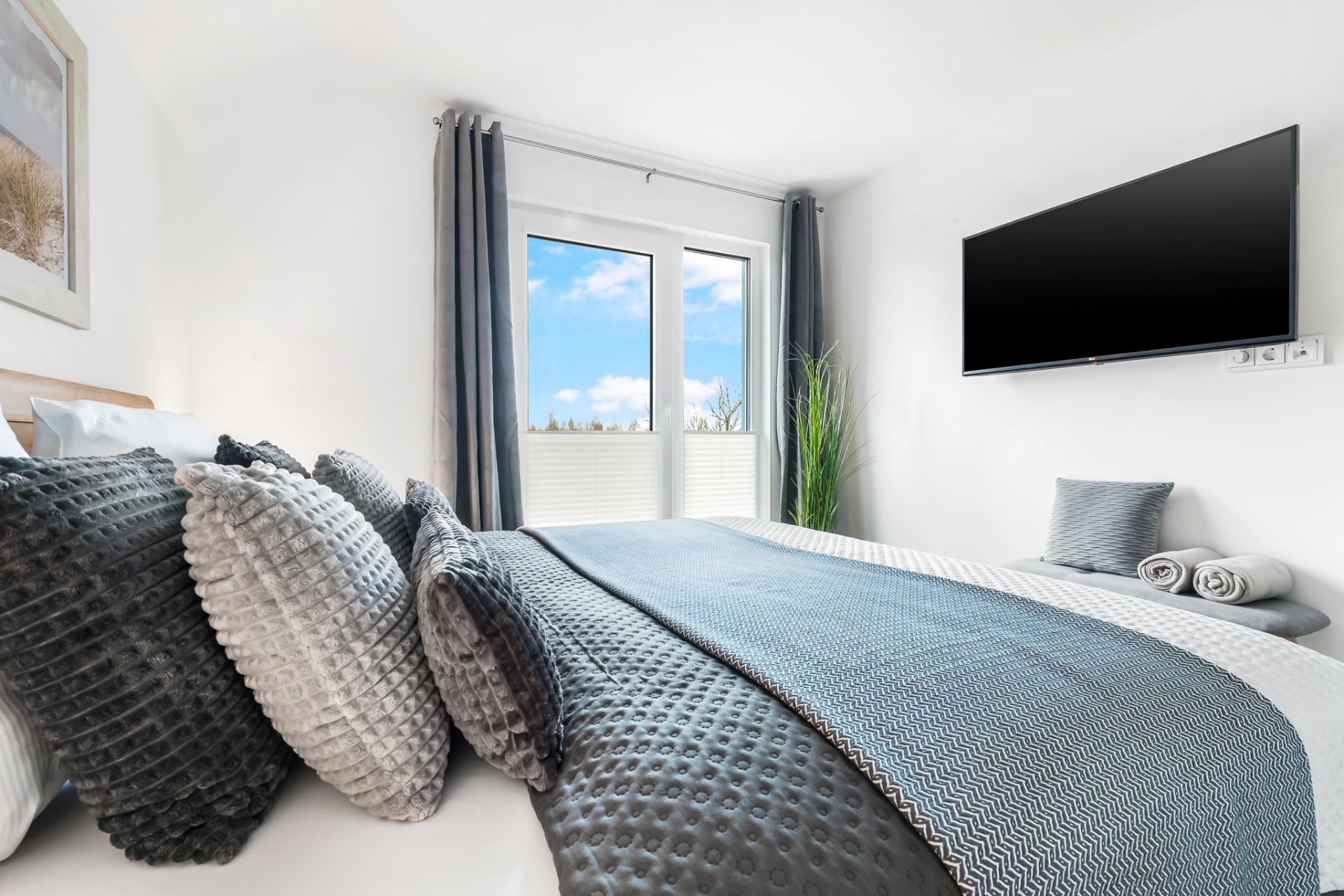 Modernes Schlafzimmer mit großem Bett, grauen Kissen, Fernseher und Fenster mit Ausblick.