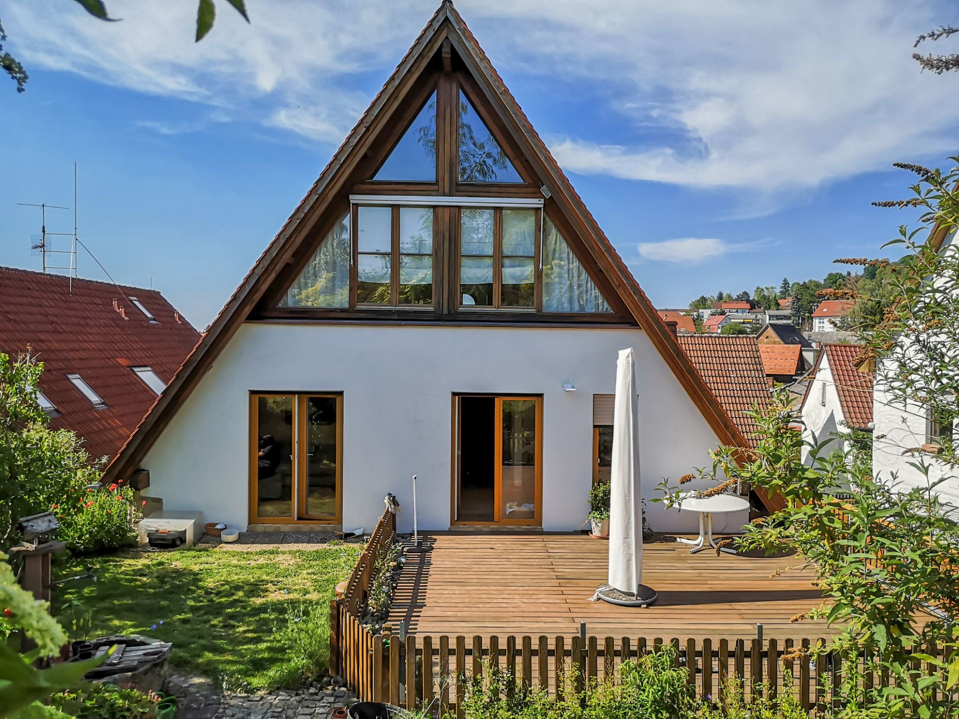 Haus mit spitzem Dach, großer Holzterrasse und Garten im Vordergrund.