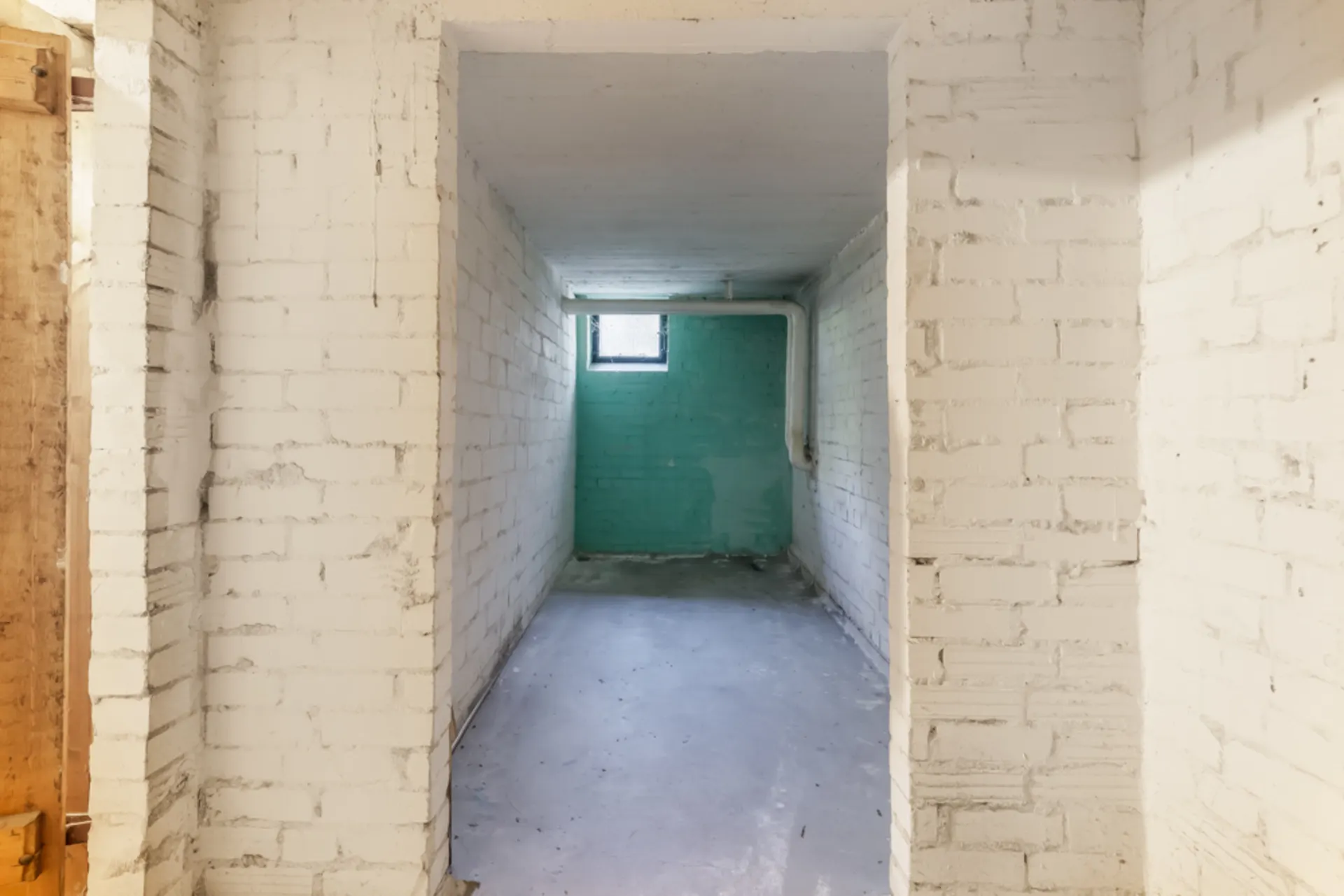Leerer Kellerraum mit weißen Backsteinwänden, kleinem Fenster und sichtbarem Rohr.