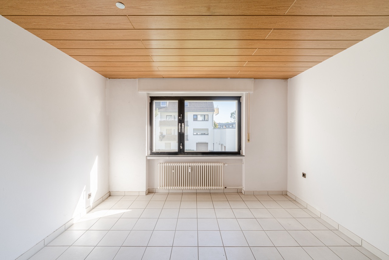 Heller leerer Raum mit Fliesenboden, Holzdecke und großem Fenster mit Heizkörper.