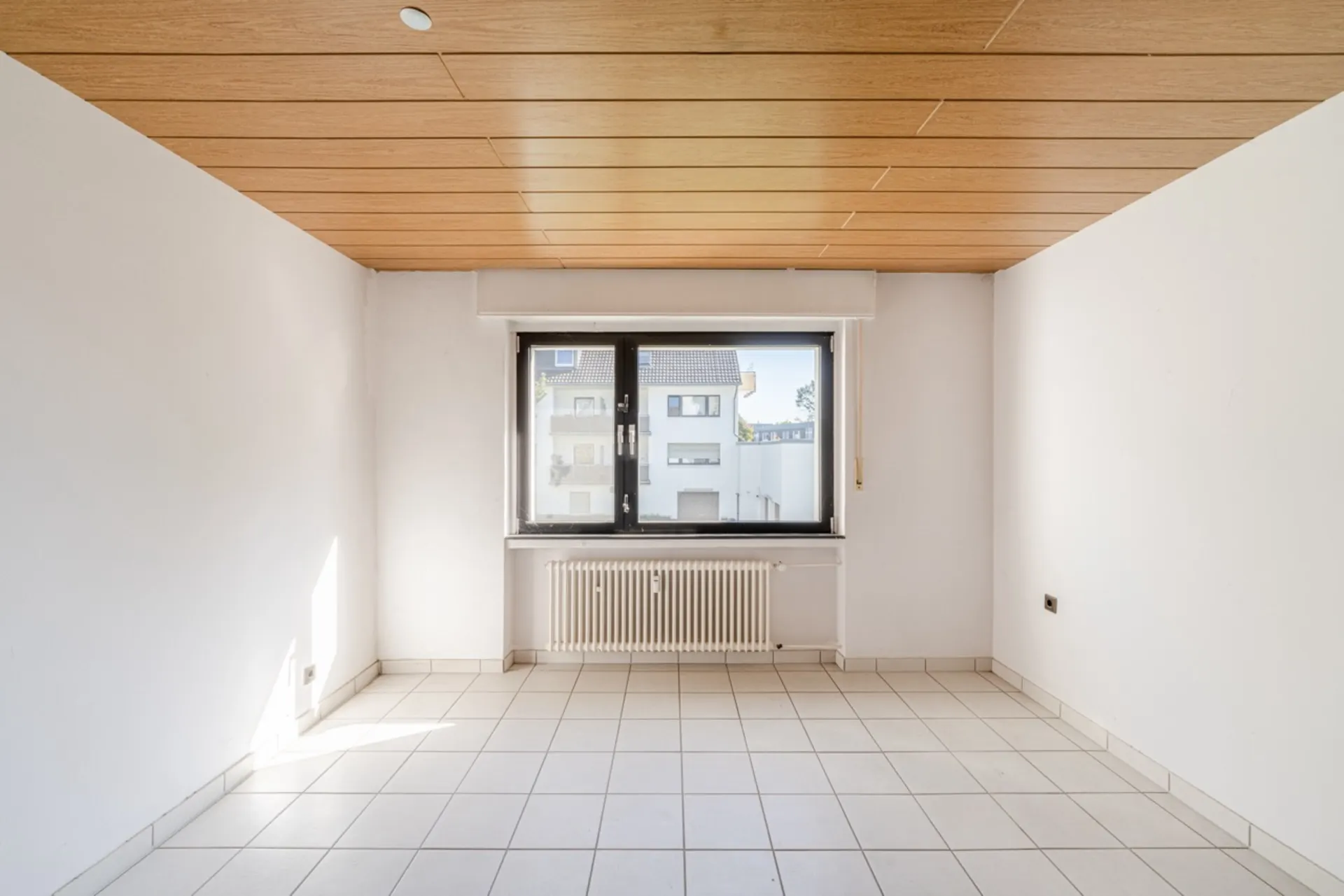Heller leerer Raum mit Fliesenboden, Holzdecke und großem Fenster mit Heizkörper.