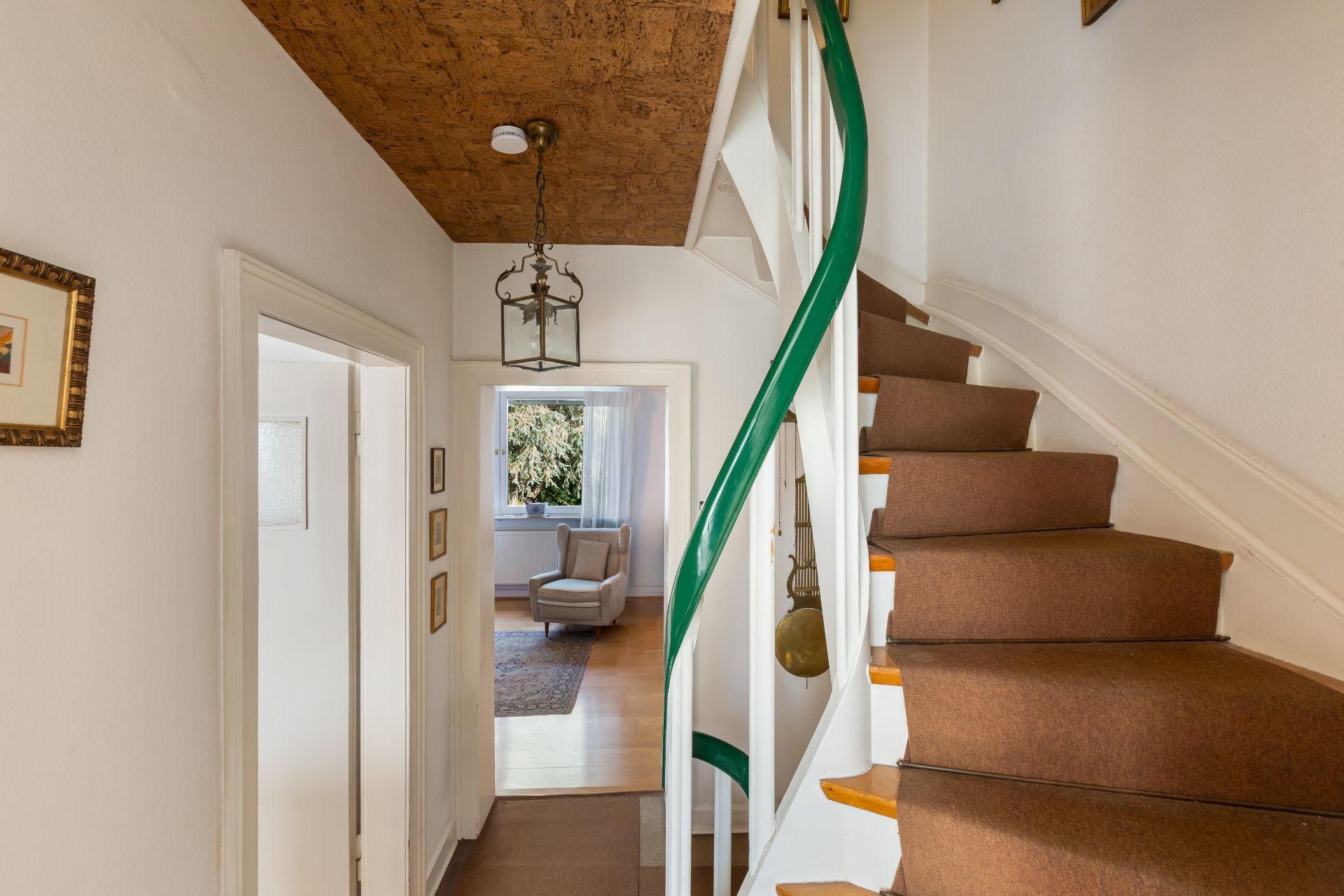 Flur mit Treppe und Blick in ein helles Wohnzimmer mit Sessel und Fenster.